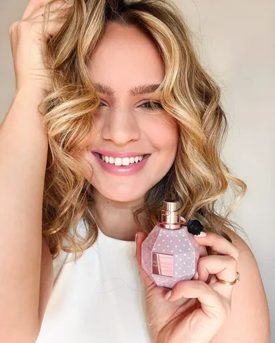 Try on this captivating wavy, blonde hairstyle showcased by a smiling woman holding a perfume bottle.