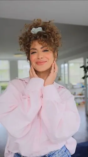 Try on a curly updo with a scrunchie accessorizing the top, as seen on a woman facing forward and smiling