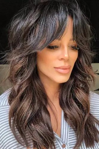 Try on this long, wavy hairstyle with bangs, featured on a woman facing forward. The hair is dark with subtle highlights enhancing the wave definition.