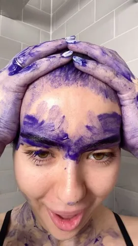 Try on this unique hairstyle moment with a hair dyeing process: a woman has her head covered in vibrant purple hair dye, hands pressing the dye into her scalp, creating an engaging and playful look.