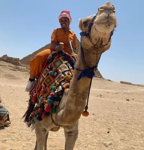 Try on the warm head covering style worn in desert climates like the one seen in this image, where an individual is dressed in a bright orange attire, seated on a vibrantly decorated camel.