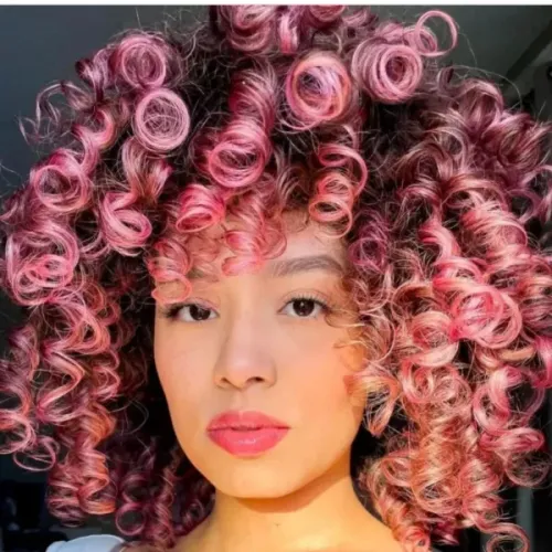 Try on this vibrant curly dyed hairstyle featuring shades of pink and dark undertones, creating a stunning voluminous look with tight, bouncy curls radiating around the face.