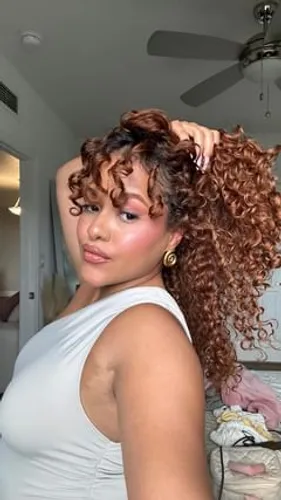 Try on this voluminous curly hairstyle, showing a young woman with thick, bouncy curls cascading down one side as she runs her fingers through them.