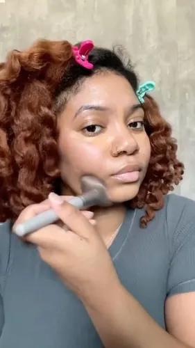 Try on this ombre hairstyle featuring voluminous curly locks in a rich auburn shade transitioning to a vibrant copper tone, held up on one side with a pink clip for a playful touch.