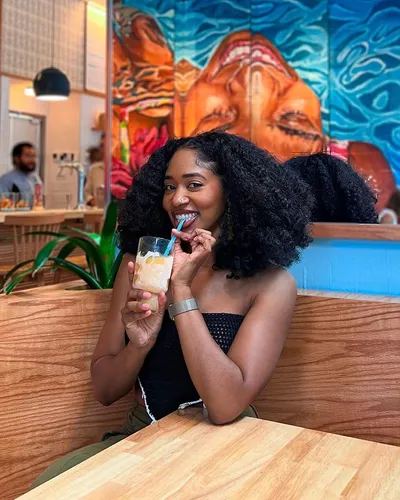Try on this voluminous shoulder-length curly hairstyle, as seen on a woman enjoying a drink at a cafe with vibrant background art.