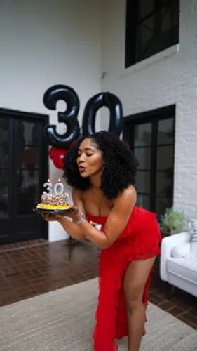 Try on this hairstyle featuring long, curly hair as the young woman holds a birthday cake in front of balloons marking a 30th birthday celebration.