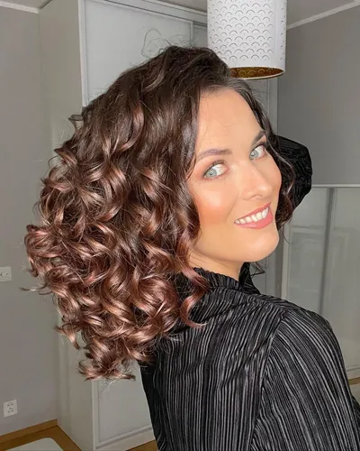 Try on this voluminous medium length curly hairstyle, with glossy brown curls cascading beautifully sideways, showcasing a lively and attractive look.