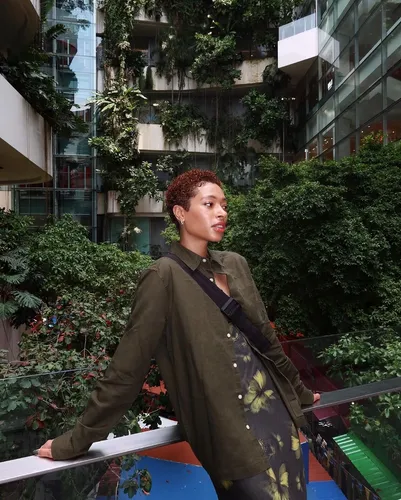 try on a hairstyle featuring short, tight curls with a warm auburn tone, presented on a woman standing in a lush, garden-like atrium.