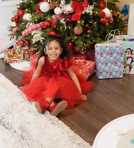 Try on this hairstyle: A young girl with a joyful smile, featuring long, curly hair tied back from her face, sitting in front of a beautifully decorated Christmas tree.