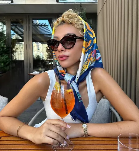 Try on a short, curly hairstyle featured on a person wearing a colorful scarf over their hair and stylish sunglasses, seated and holding a cocktail.