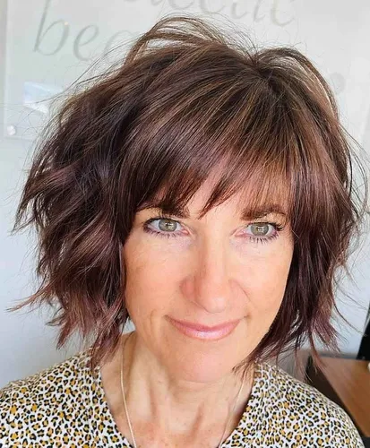 Try on this stylish wavy and tousled hairstyle with a casual fringe to frame the face. The hair color blends dark brown with subtle highlights, providing a rich texture.