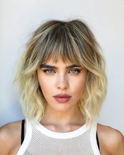 Try on this shaggy bob with light bangs, styled in loose waves and featuring a subtle balayage color transition from dark roots to lighter ends.